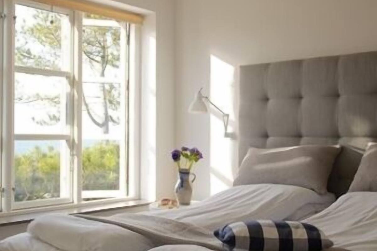 A bedroom with a large window, a bed with white and striped bedding, and a vase with flowers on the nightstand.