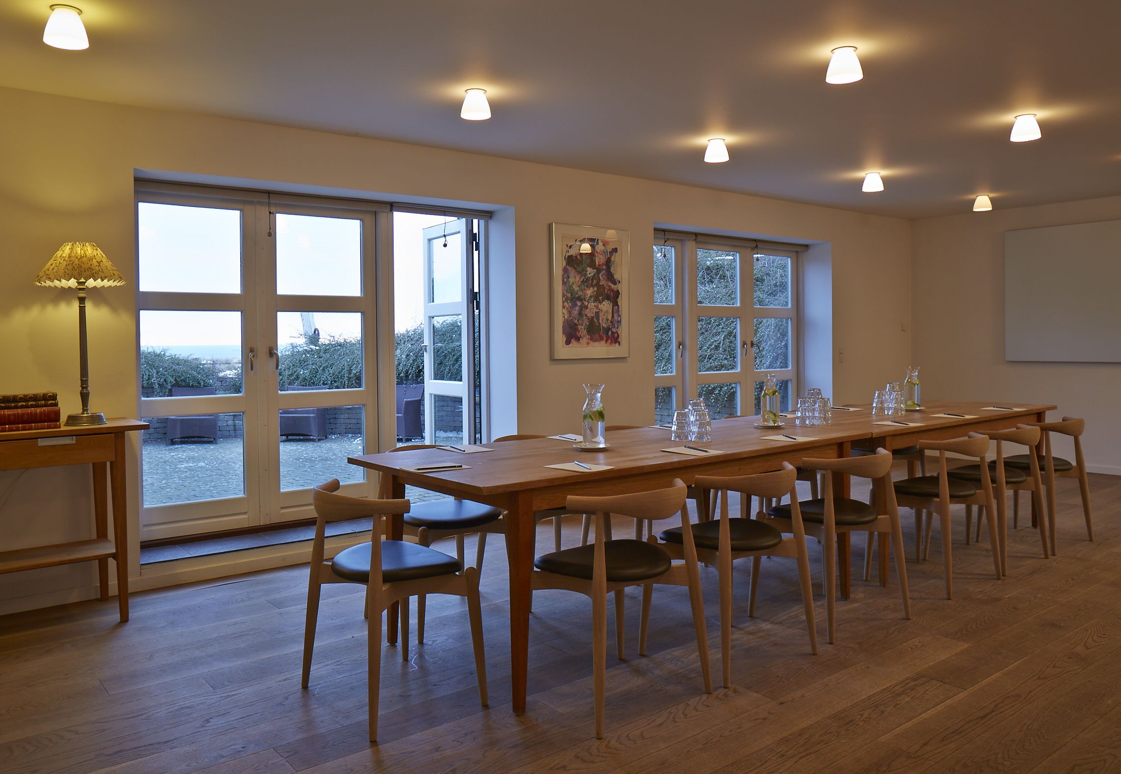 A spacious room with a large wooden table set for dining, multiple chairs, and large windows offering a view of the outdoors.