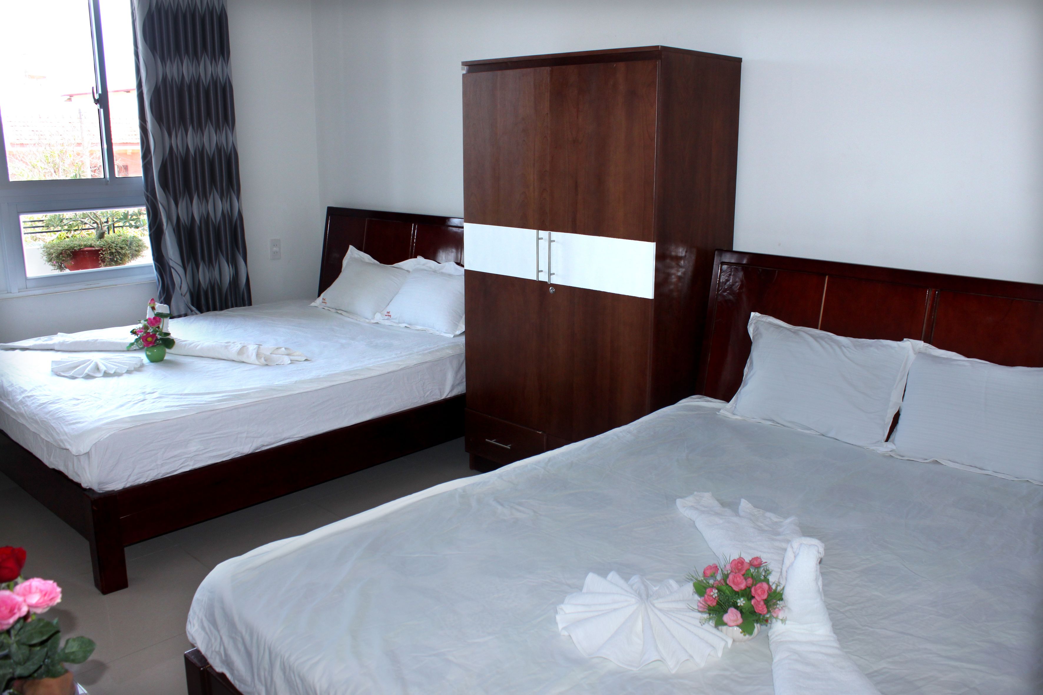 Two single beds with white linens, a wooden wardrobe, and a window with a view of a building and a potted plant.