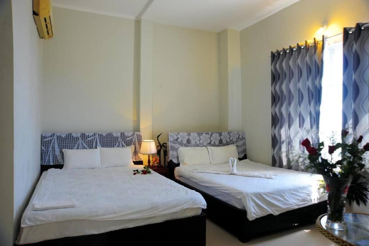 Hotellihuone, jossa on kaksi sänkyä, yöpöytä lampun kanssa, maljakko kukkineen ja ikkuna, jossa on verhot.