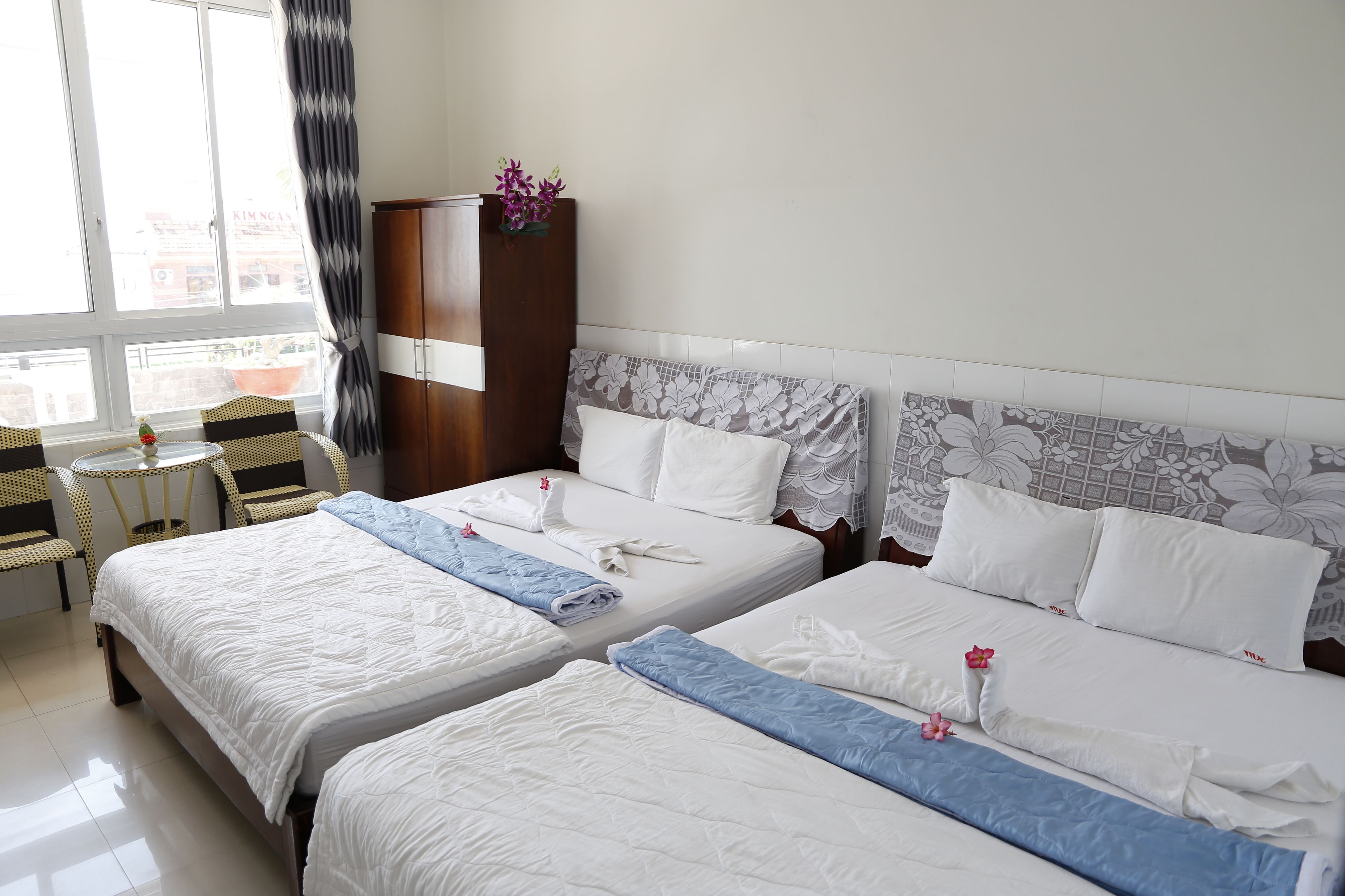 A hotel room with two beds, a wooden wardrobe, a small table with chairs, and a window with curtains.