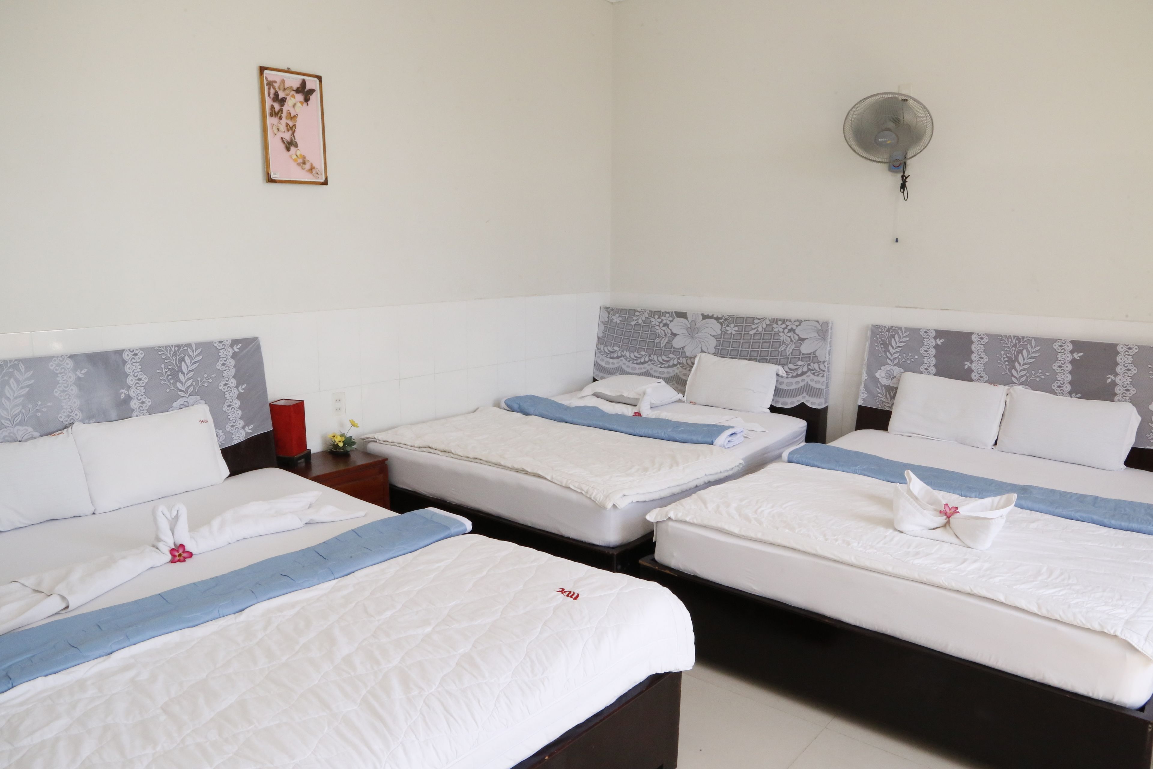 Una camera d'albergo con tre letti, ognuno con biancheria bianca e un tocco di blu. C'è una ventola a muro e un quadro appeso al muro.