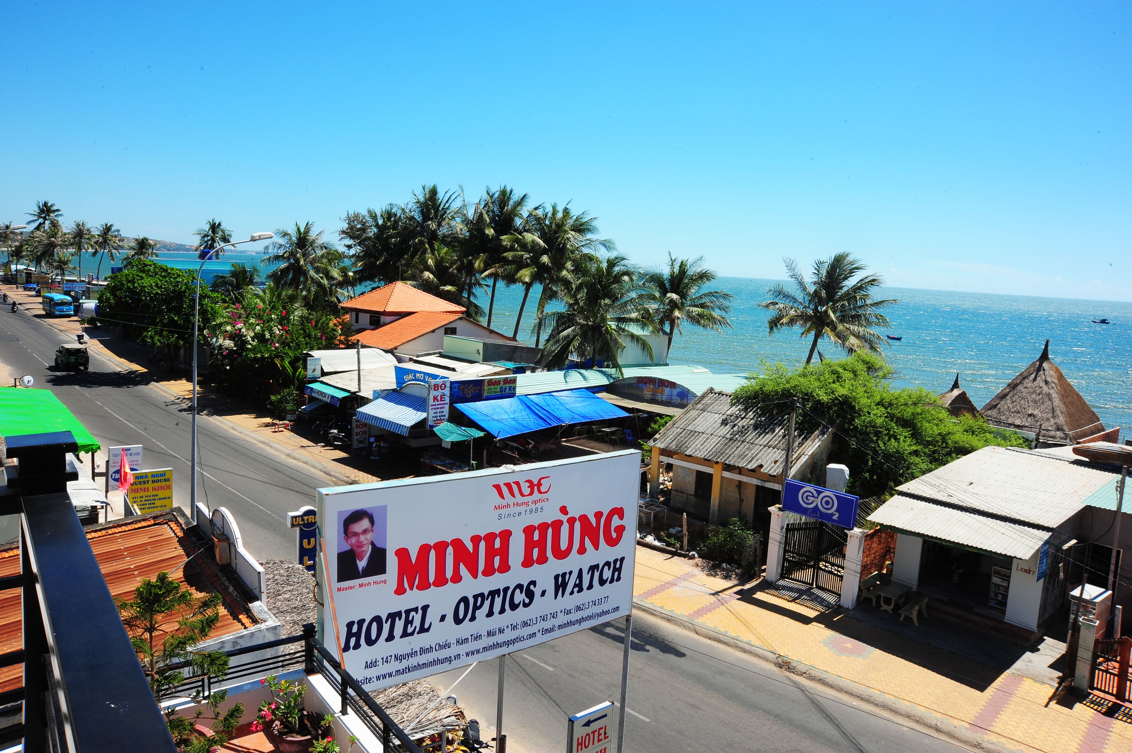 A hotel named Minh Hung with a signboard, located near a beach with palm trees and a clear blue sky.