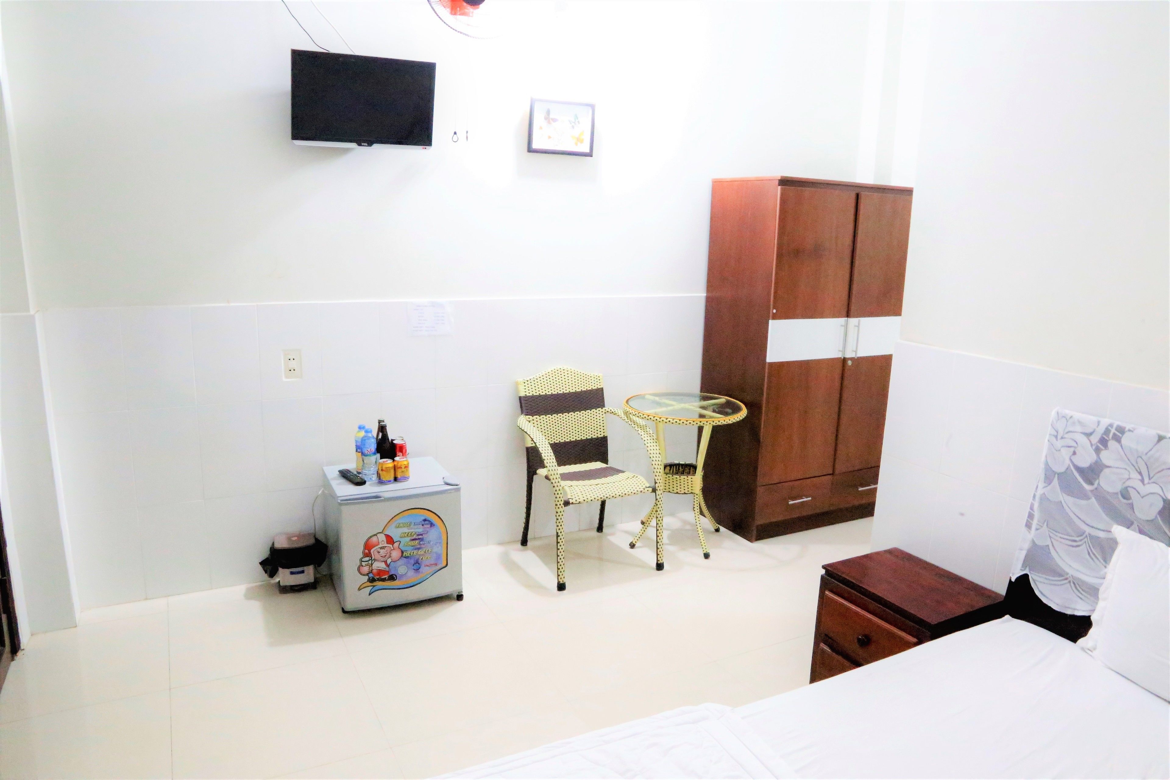 A hotel room with a bed, a wardrobe, a small table with chairs, a television on the wall, and a small cabinet with items on it.
