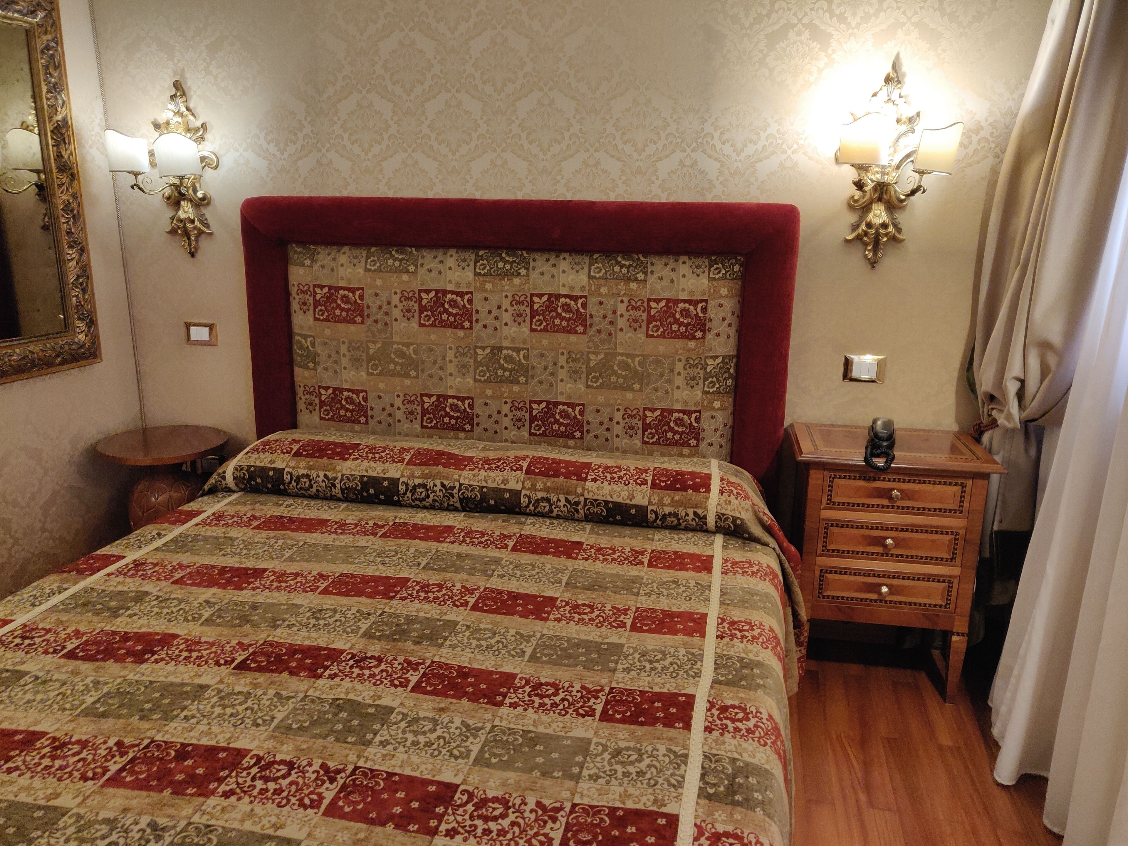 Un lit bien fait, avec une tête de lit rouge, une table de chevet en bois dotée de tiroirs, un miroir et deux appliques murales.