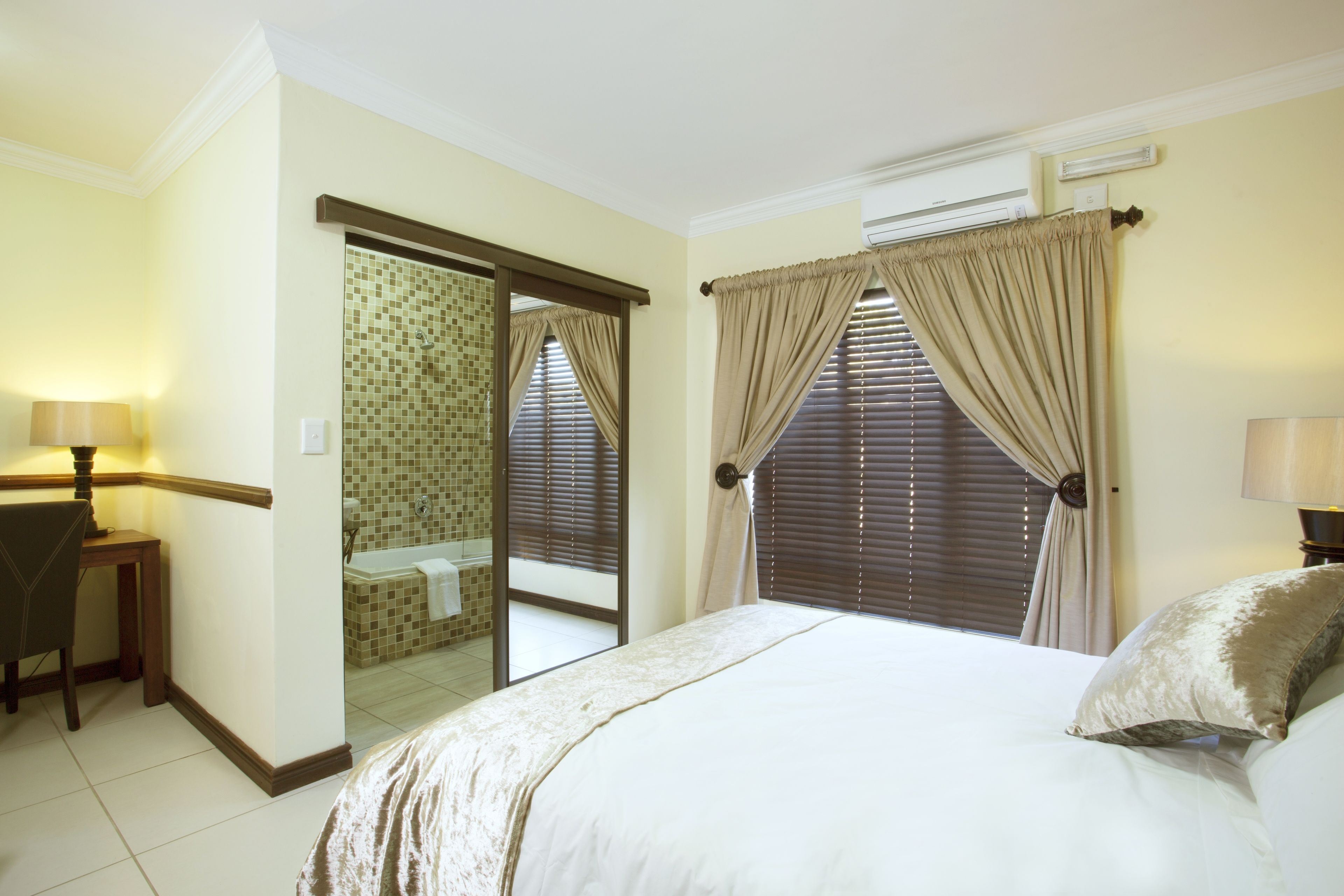 Habitación de hotel con cama, escritorio, espejo y baño visible a través de una puerta abierta.