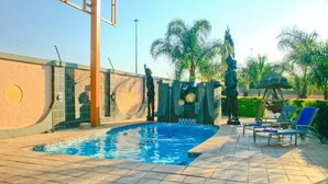 Una piscina con borde de baldosas, sillas de descanso y una estatua.