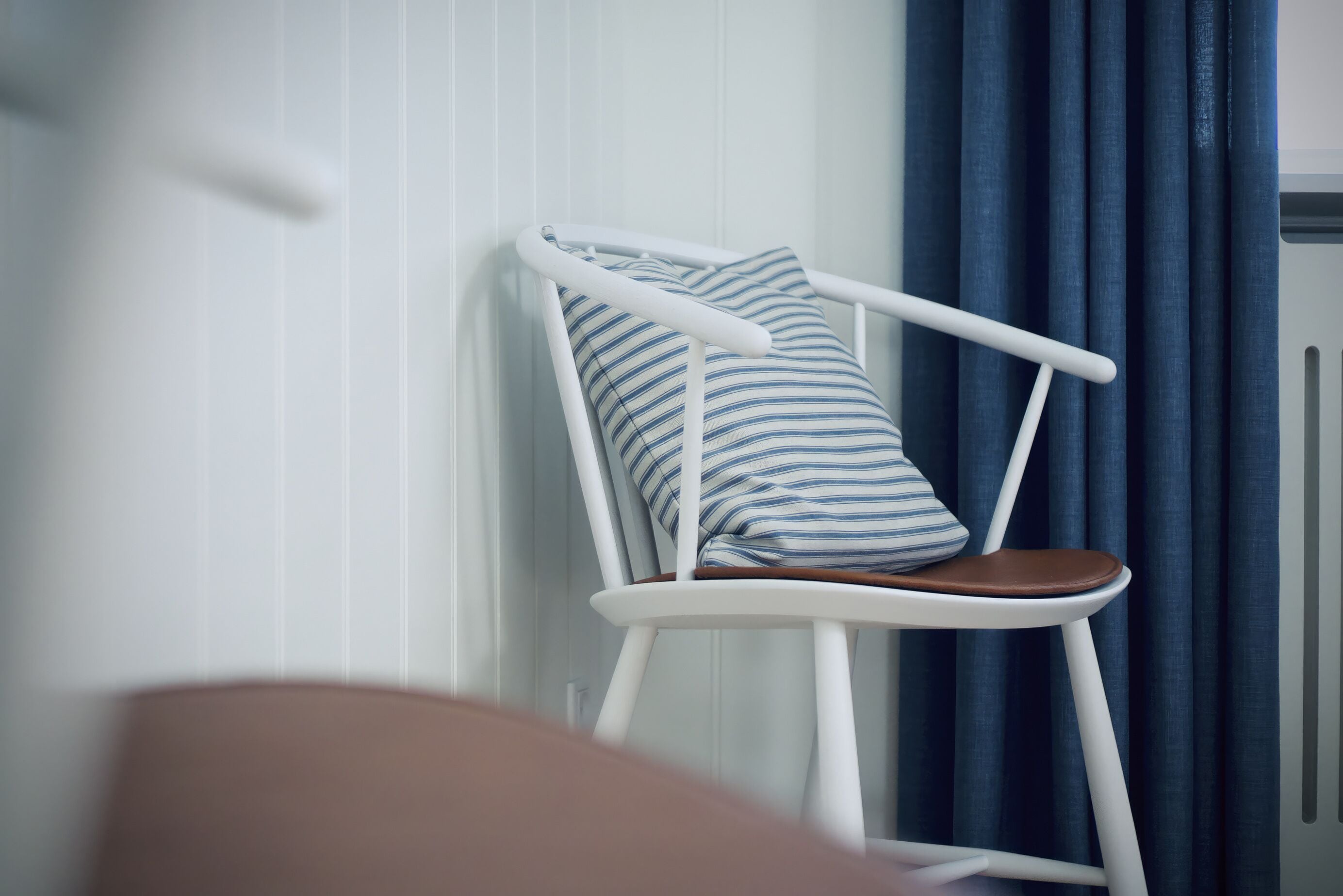 A white chair with a striped pillow against a white wall, next to a blue curtain.