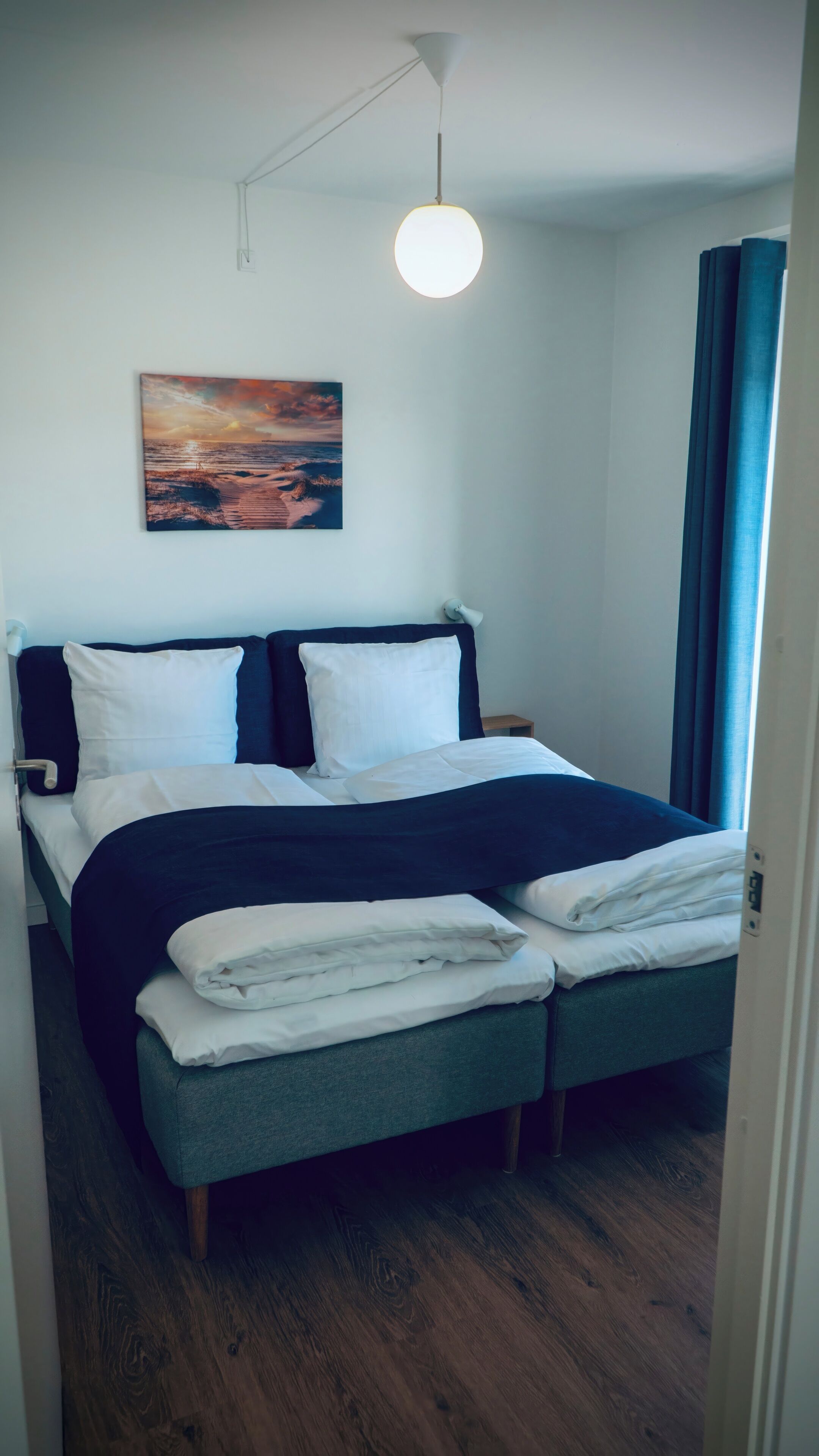 A neatly made bed with white and dark bedding, a wall-mounted painting of a sunset, and a hanging light fixture.