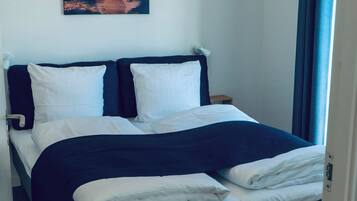 A neatly made bed with white and dark bedding, a wall-mounted painting of a sunset, and a hanging light fixture.