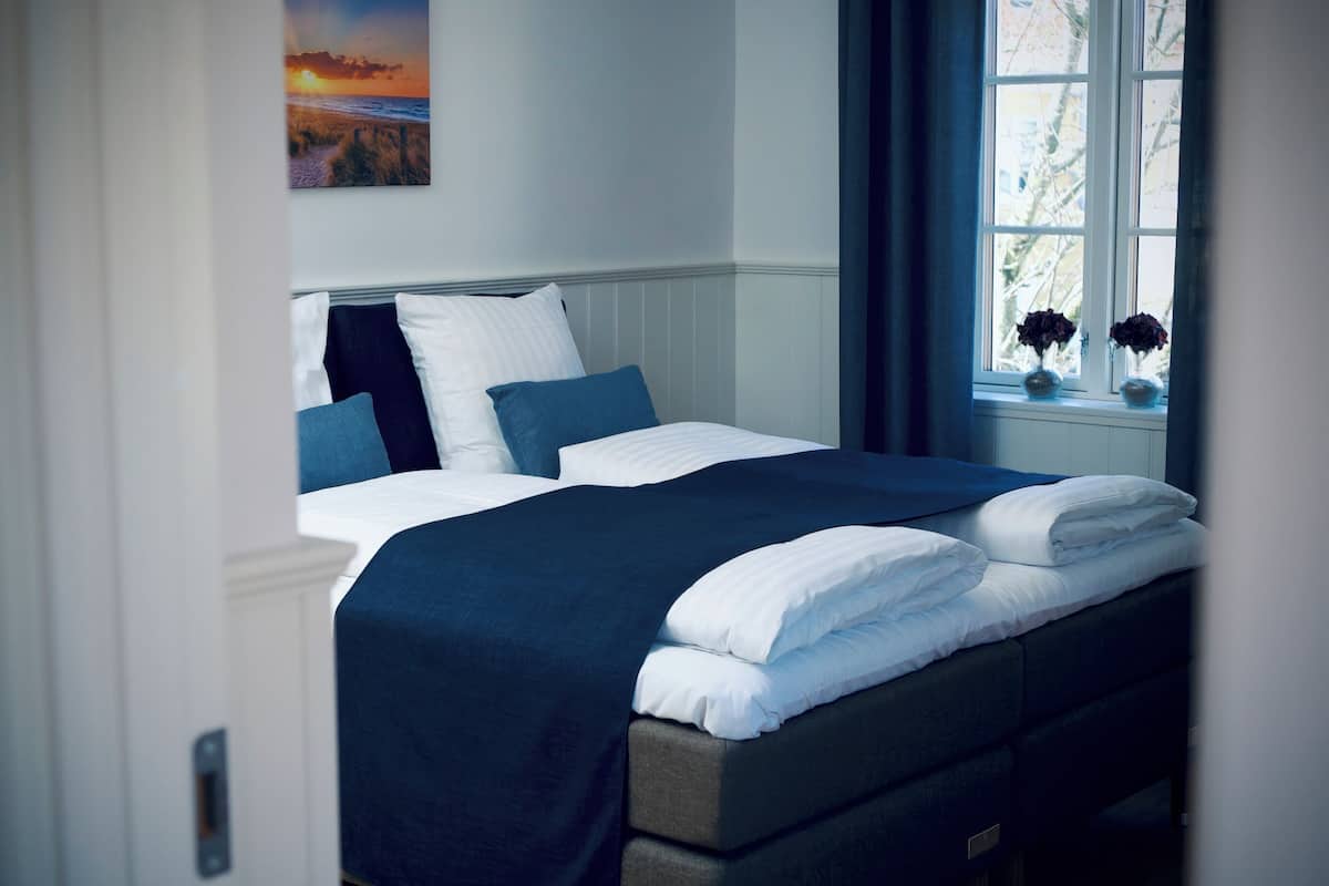 A neatly made bed with white and blue bedding, a window with a view of trees, and a painting of a sunset on the wall.