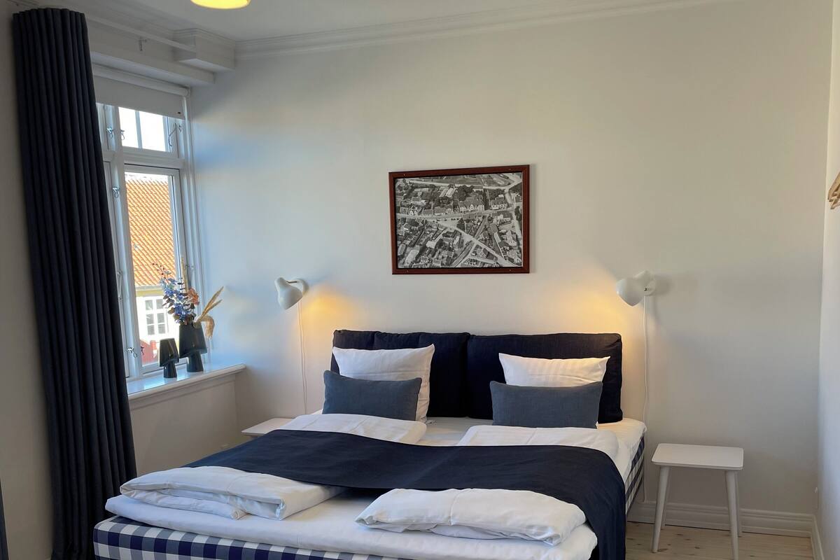 A bedroom with a checkered bedspread, a window with curtains, a framed picture on the wall, and a small white stool.
