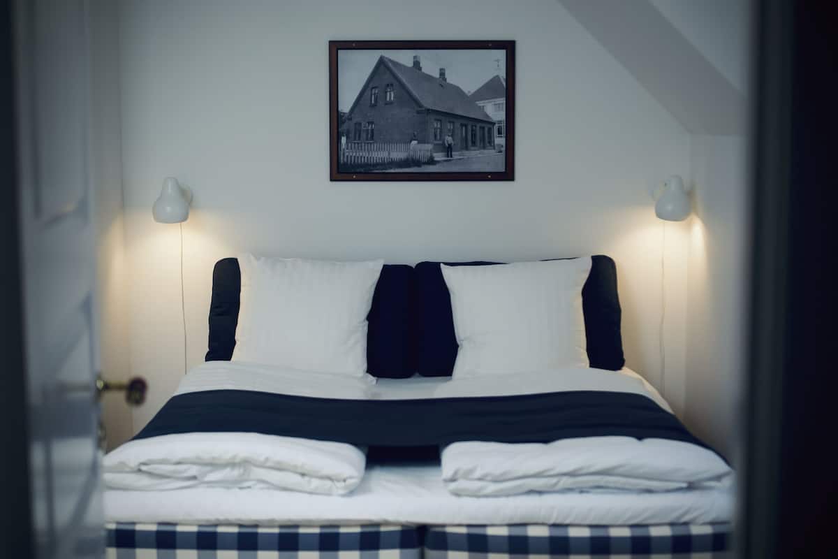 A neatly made bed with white and dark bedding, a framed picture of a house on the wall, and two wall-mounted lamps.