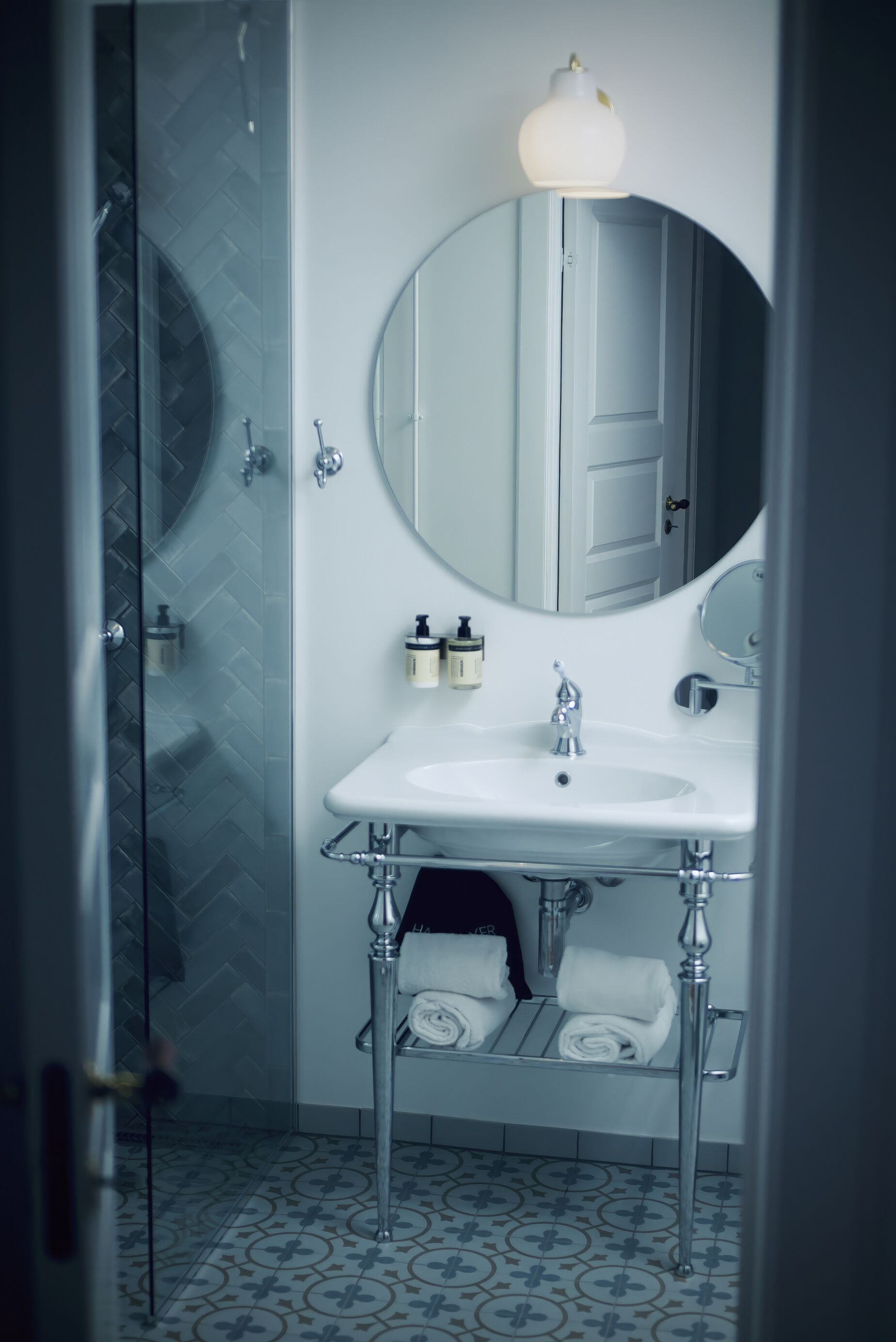 A modern bathroom with a glass shower enclosure, a white sink with a round mirror, and a towel rack.