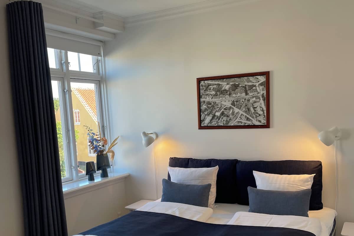 A neatly made bed with a checkered bedspread, a framed black and white photograph on the wall, and a window with curtains.