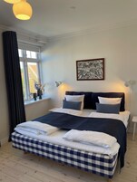 Una cama bien tendida con una colcha a cuadros, una fotografía en blanco y negro enmarcada en la pared y una ventana con cortinas.