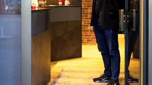 A man standing in a modern lobby with a brick wall and a reception desk. - Hotel Ambassador Essen (Essen)