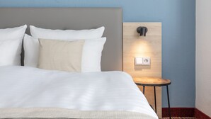 A neatly made bed with white linens and a wooden headboard. A small wooden nightstand with a wall-mounted lamp is beside the bed. - Hotel Ambassador Essen (Essen)