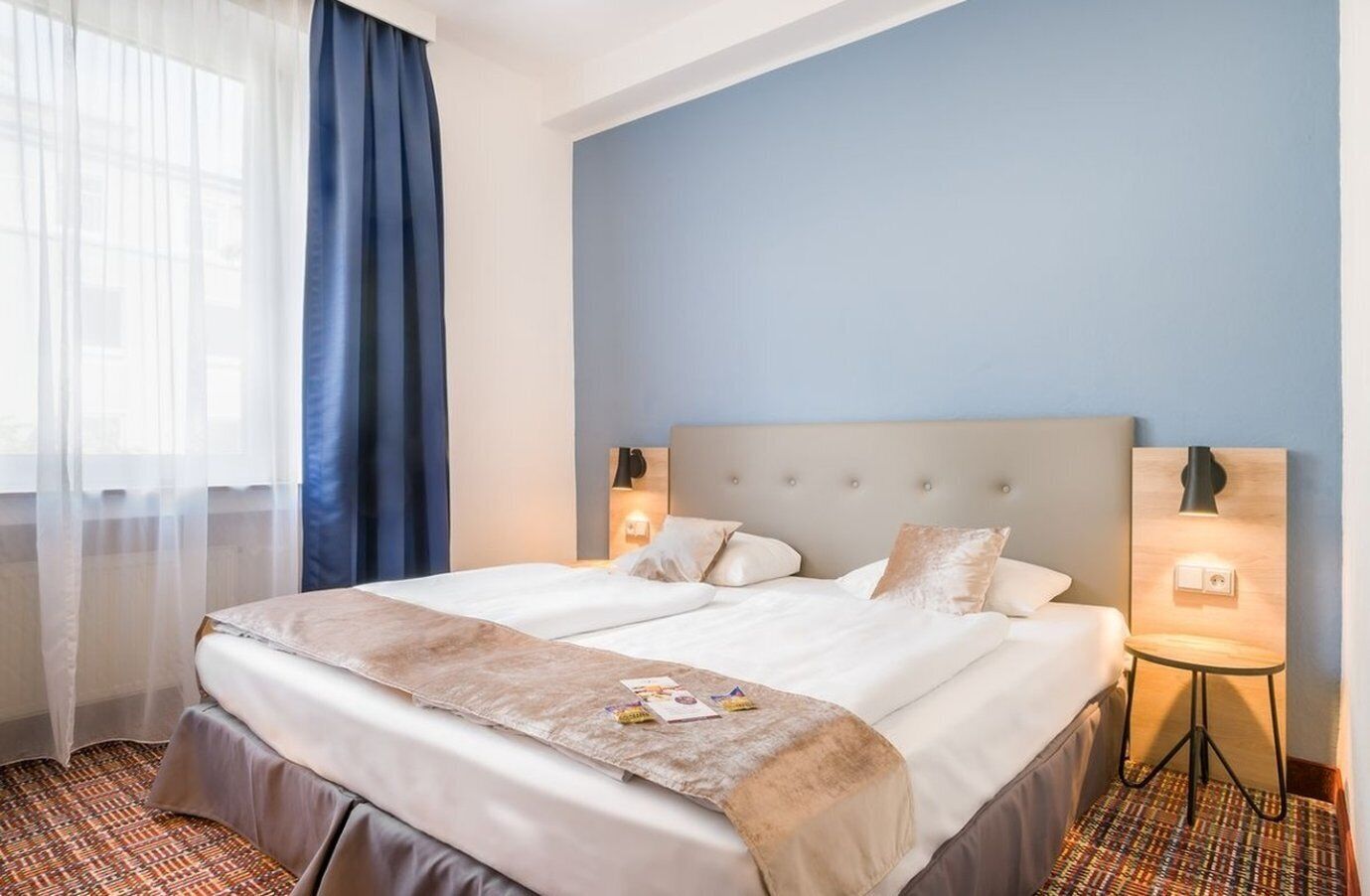 A hotel room with a large bed, a headboard with a light fixture, a bedside table with a lamp, and a window with sheer curtains.