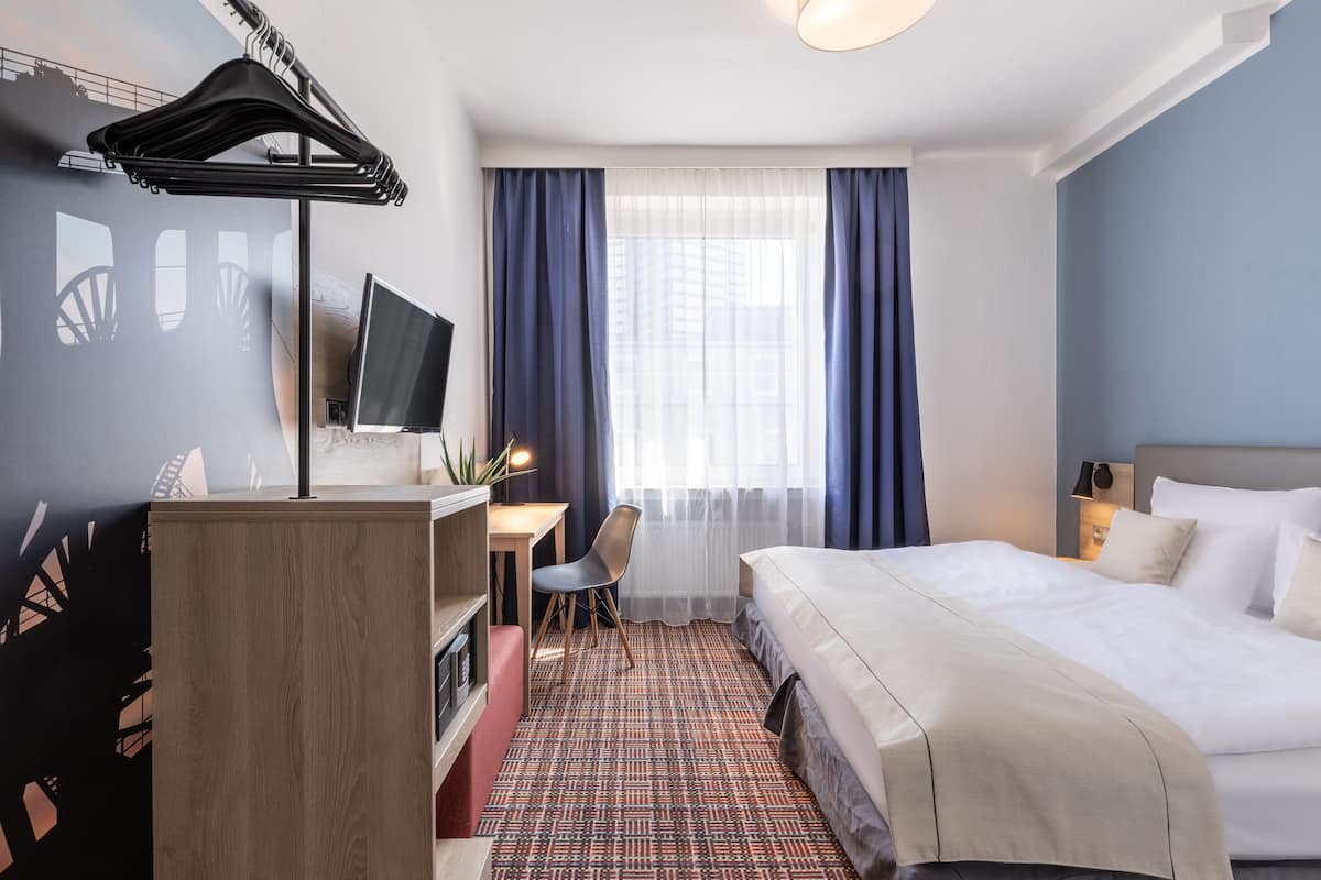 A hotel room with a bed, desk, chair, TV, and a view of the city through the window.