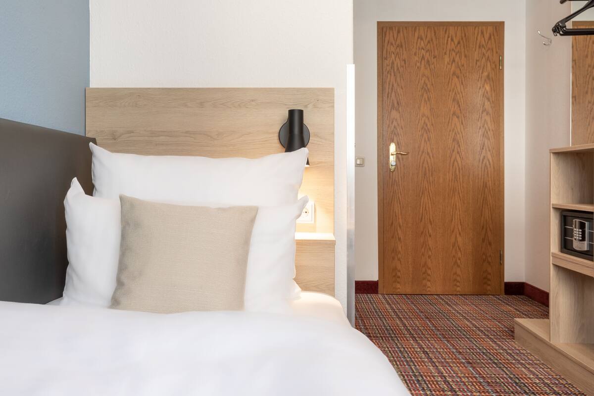 A hotel room with a bed, a wooden headboard, a nightstand with a lamp, a door, and a patterned carpet.