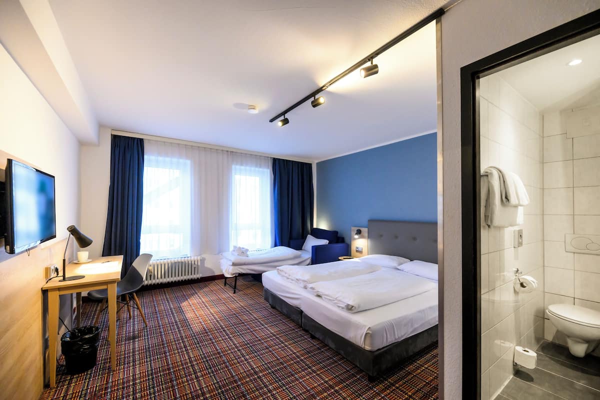 A hotel room with two beds, a desk, a chair, a TV, and a bathroom with a toilet and shower.