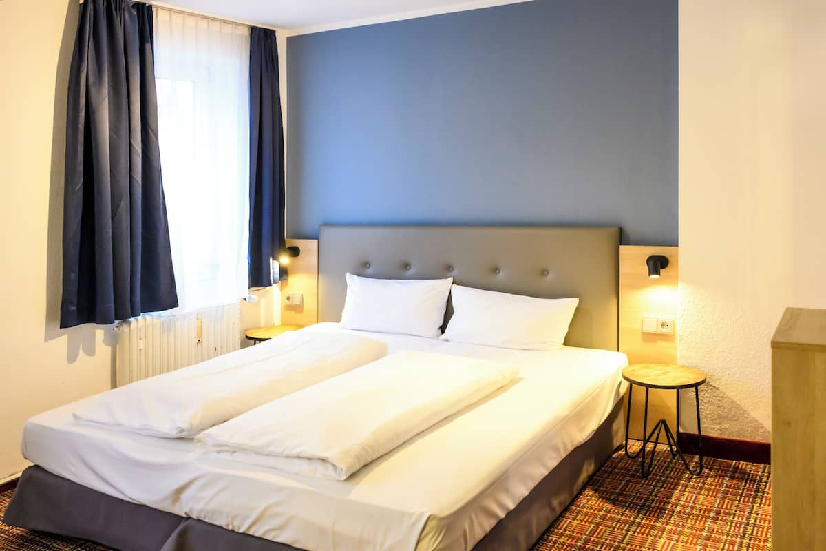 A neatly made double bed with white linens, a headboard, two bedside lamps, and a patterned carpet.