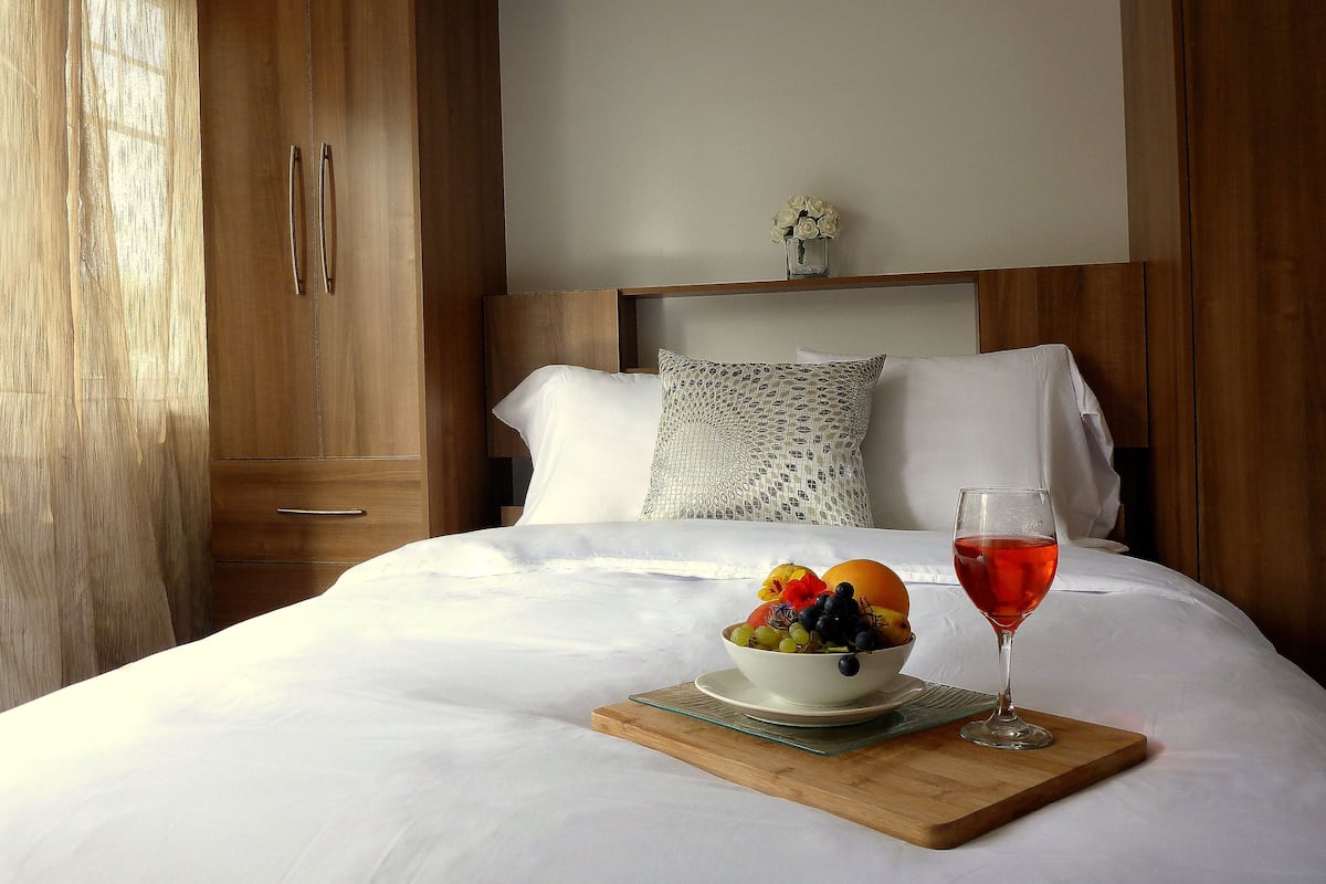 Una cama bien hecha con sábanas blancas, un cuenco de fruta y un vaso de bebida roja sobre una bandeja de madera.
