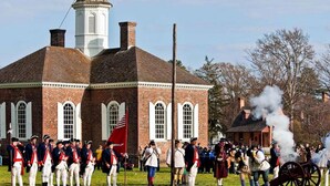 A historical reenactment with participants in period costumes, a cannon firing, and a brick building with a white tower. - Bassett Motel (Williamsburg)