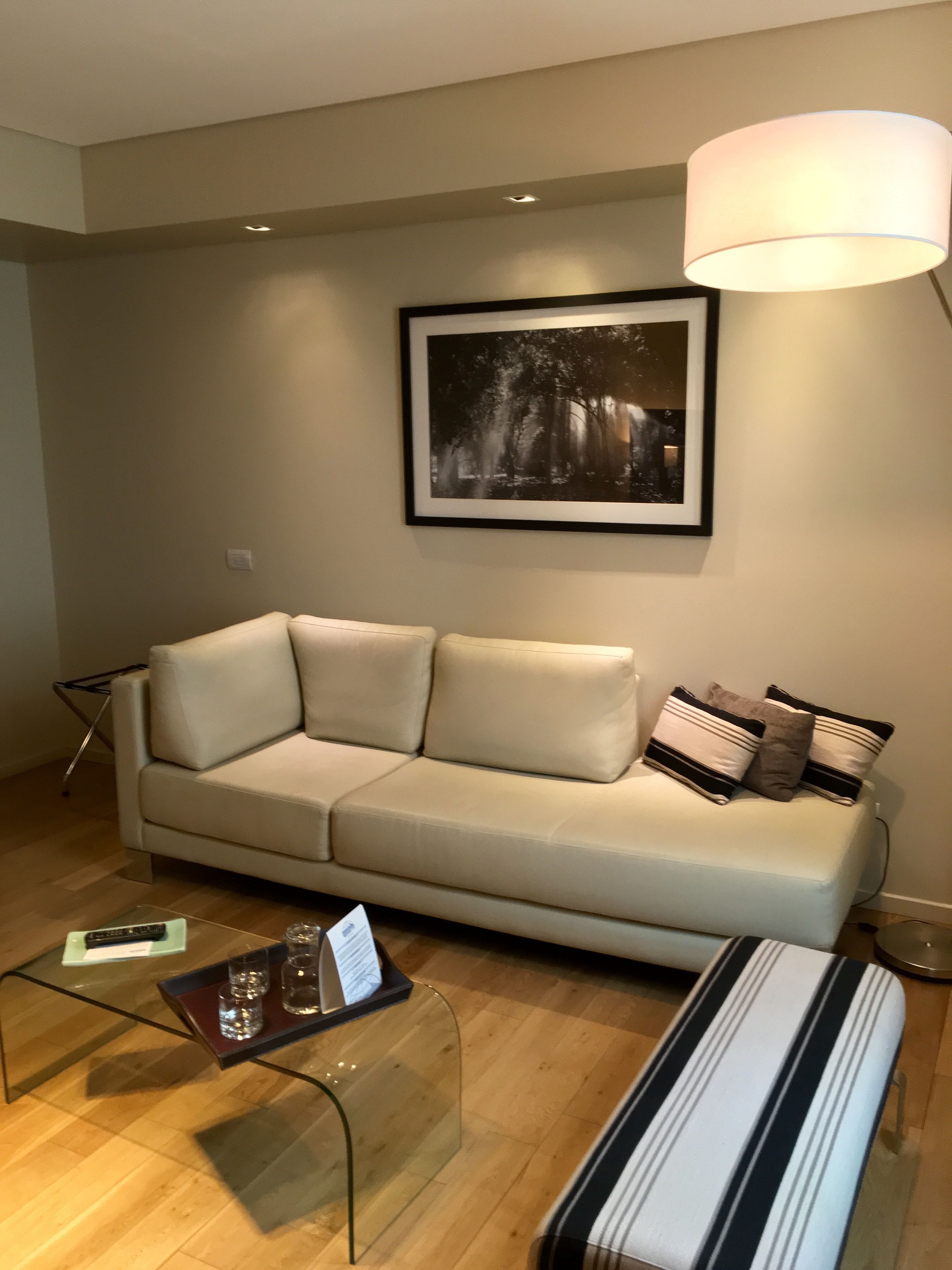 Uma sala de estar com um sofá bege, uma mesa de centro de vidro com controle remoto e copos, um quadro na parede e um abajur.