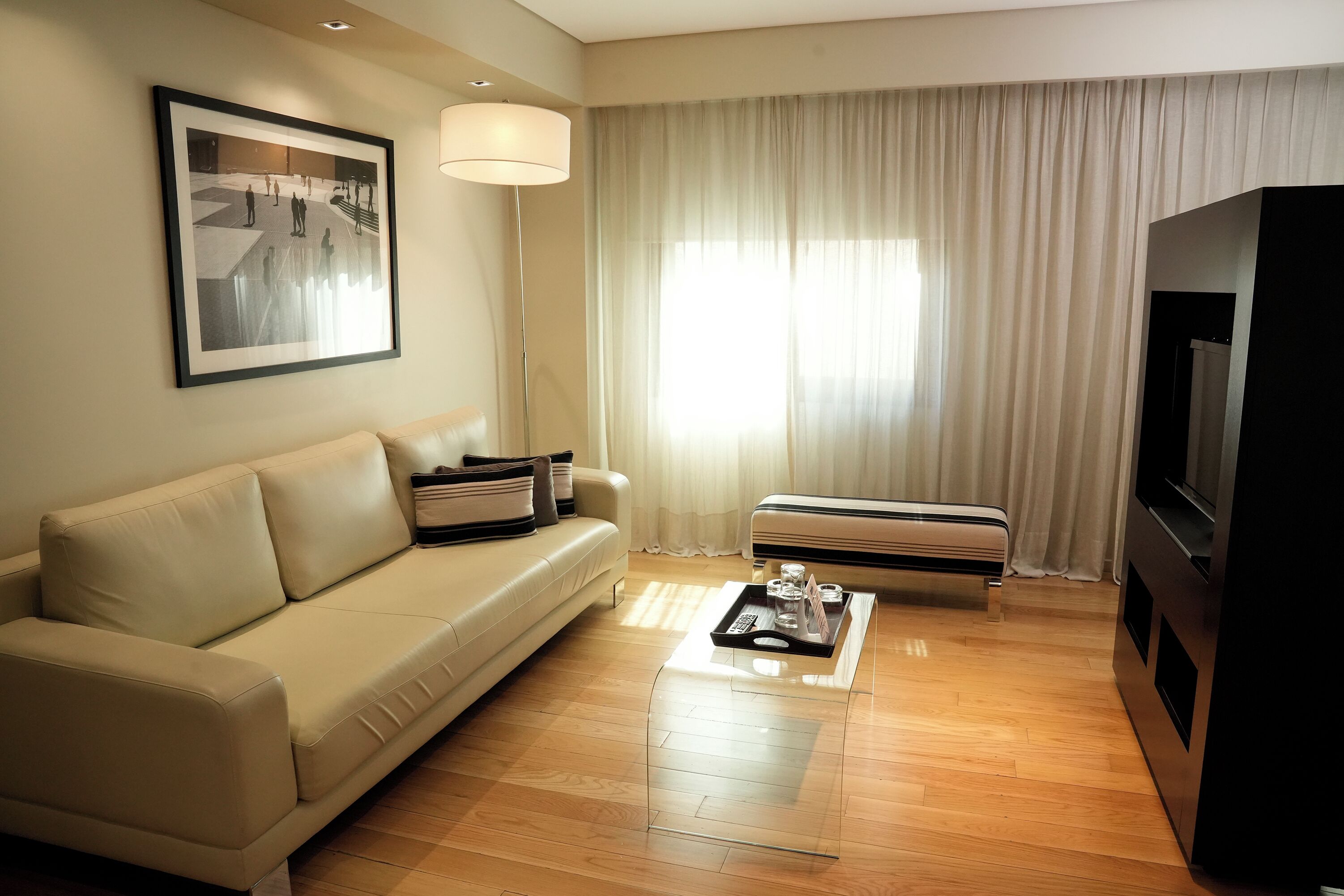 Una sala de estar moderna con un sofá beige, una mesa de centro con una bandeja, un televisor de pantalla plana y un ventanal con cortinas.
