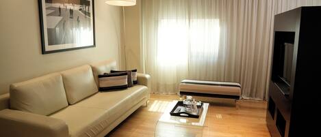 Una sala de estar moderna con un sofá beige, una mesa de centro con una bandeja, un televisor de pantalla plana y un ventanal con cortinas.