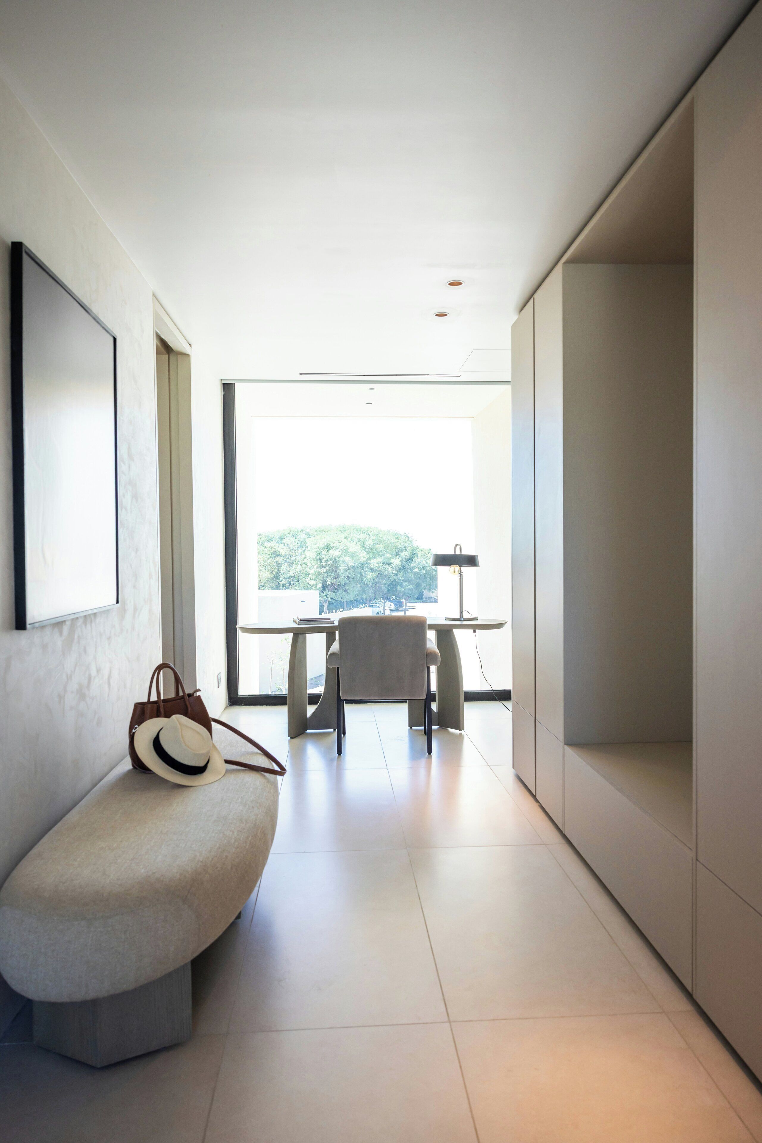 Un pasillo moderno y minimalista con un banco, una mesa con una lámpara y vistas al exterior con vegetación.