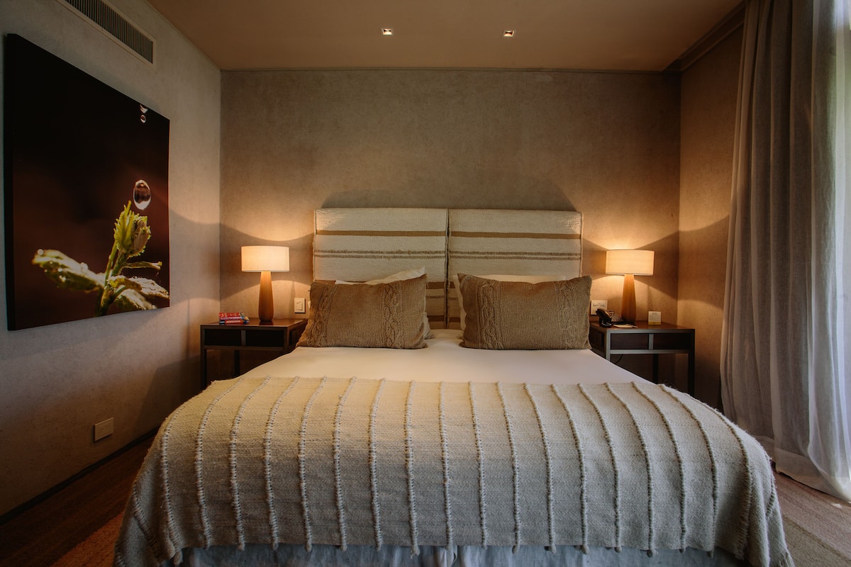 A hotel room with a large bed, two bedside lamps, a headboard, and a painting on the wall.