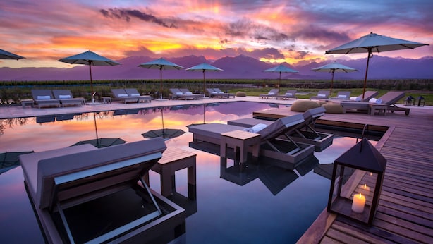 Un área de piscina tranquila con sillones reclinables y sombrillas, rodeada por una terraza de madera y que refleja una puesta de sol sobre montañas.