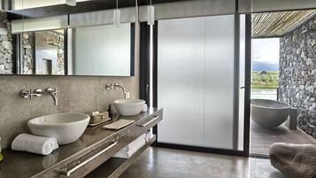 A modern bathroom with a stone wall, two sinks, a large bathtub, and a view of the outdoors.