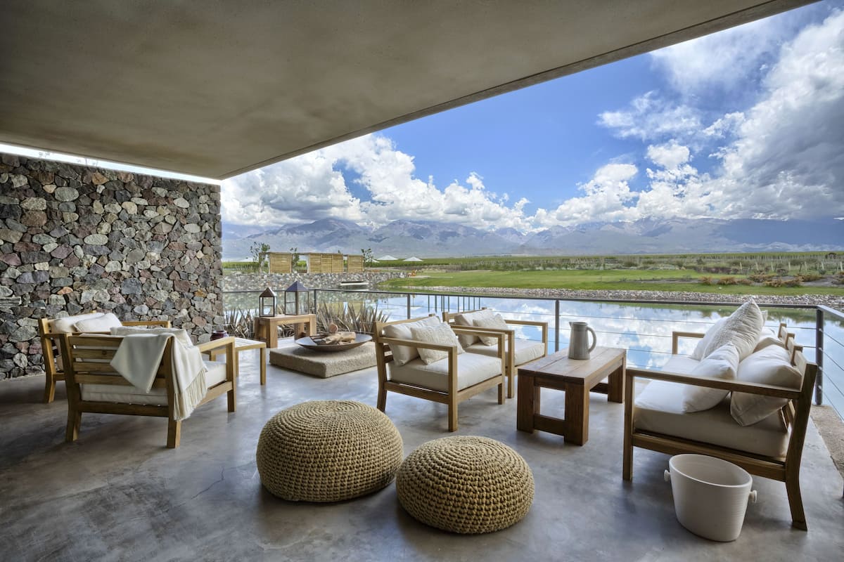 A spacious outdoor seating area with stone walls, wooden furniture, and a view of mountains and a lake.