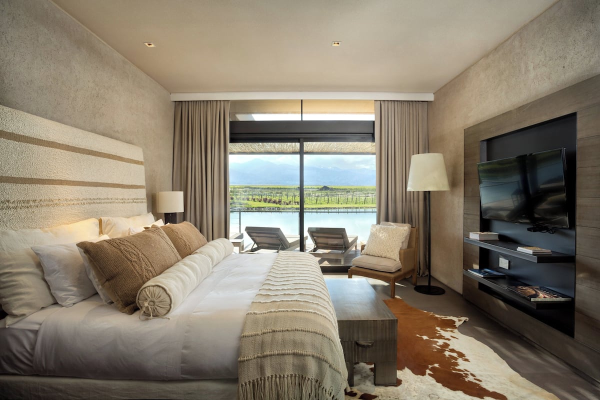 A modern hotel room with a large bed, two armchairs, a TV, and a view of a lake and mountains.