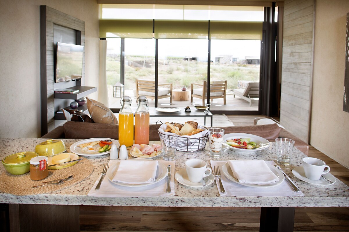 Un desayuno con una mesa dispuesta para dos personas, incluyendo platos, cubiertos y una cesta de pan.