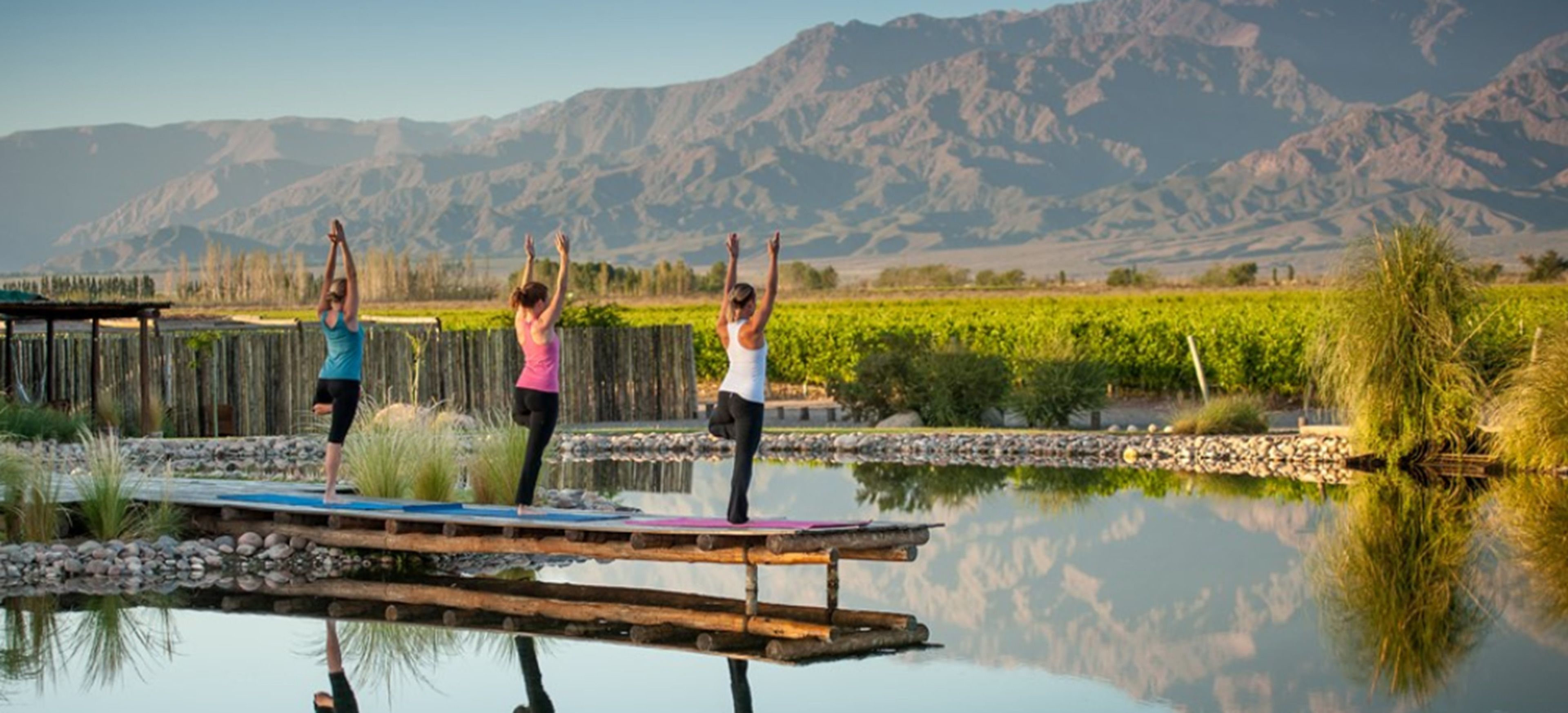 Three people practicing yoga on a wooden platform over a calm body of water, with a vineyard and mountains in the background.