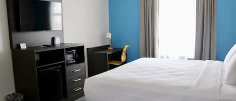 Habitación de hotel con una cama grande, un televisor de pantalla plana en la pared, un escritorio con lámpara y vistas al exterior a través de una ventana con cortinas.