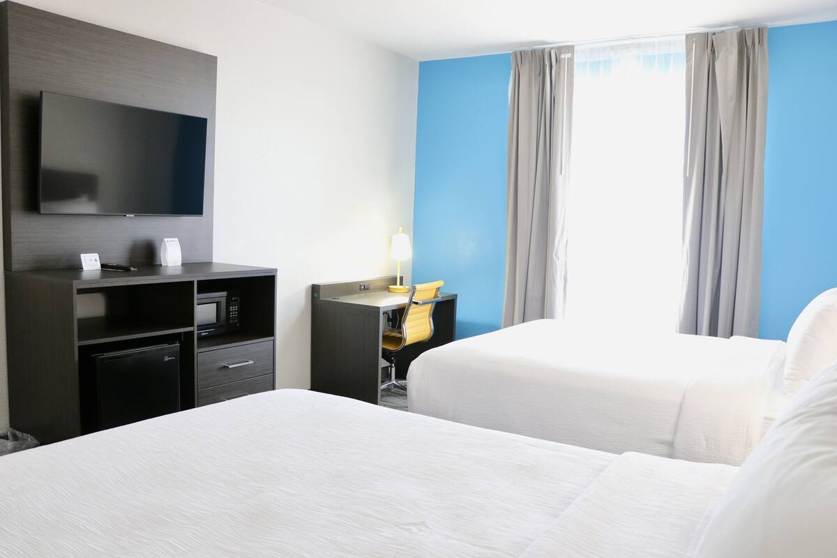 A hotel room with a flat-screen TV, a desk with a chair, a bed, and a window with curtains.