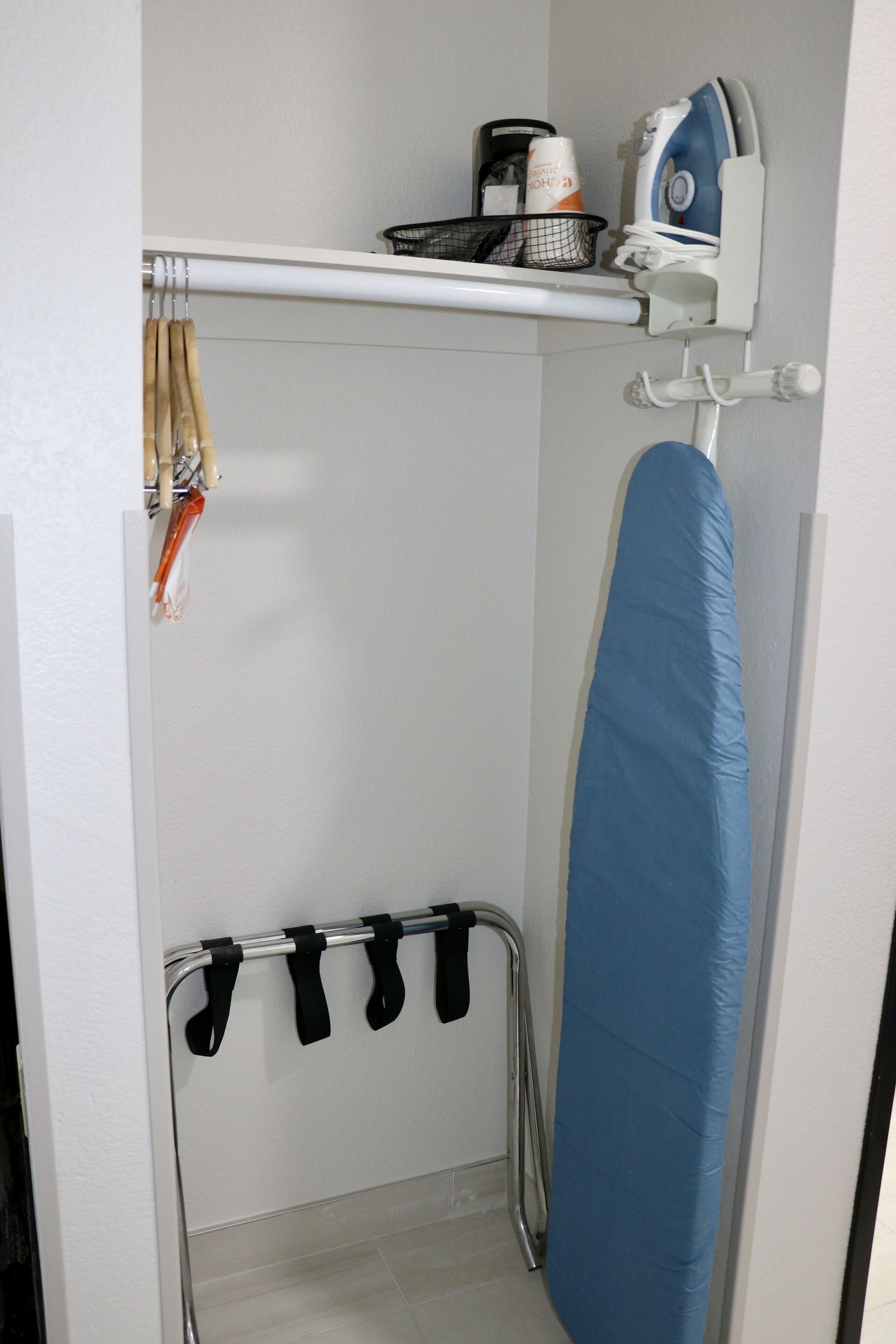 A small laundry area with an ironing board, a drying rack, and a shelf with a coffee maker and other items.