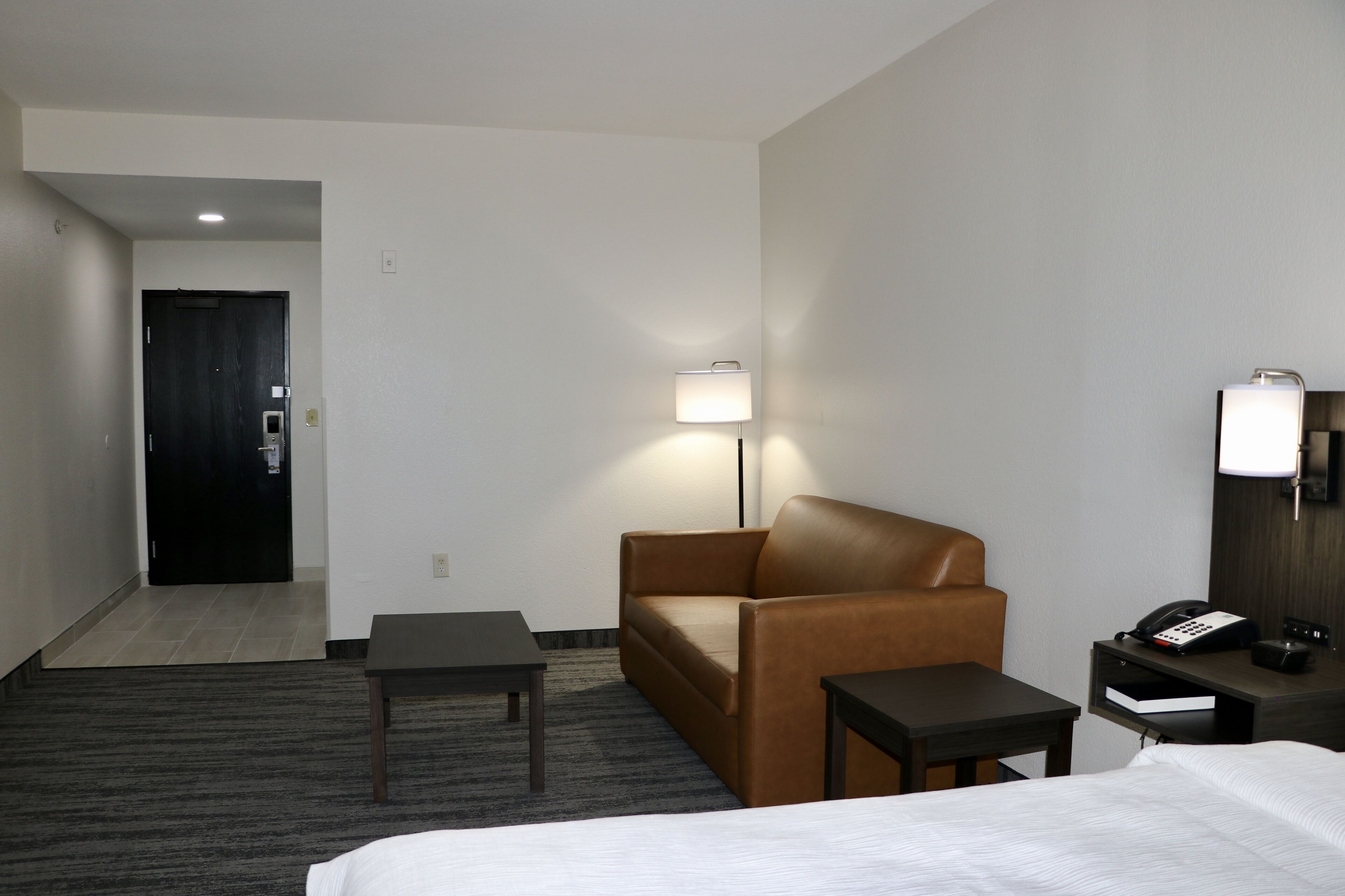 A hotel room with a bed, a brown leather armchair, a small table, a nightstand with a phone, and a lamp.