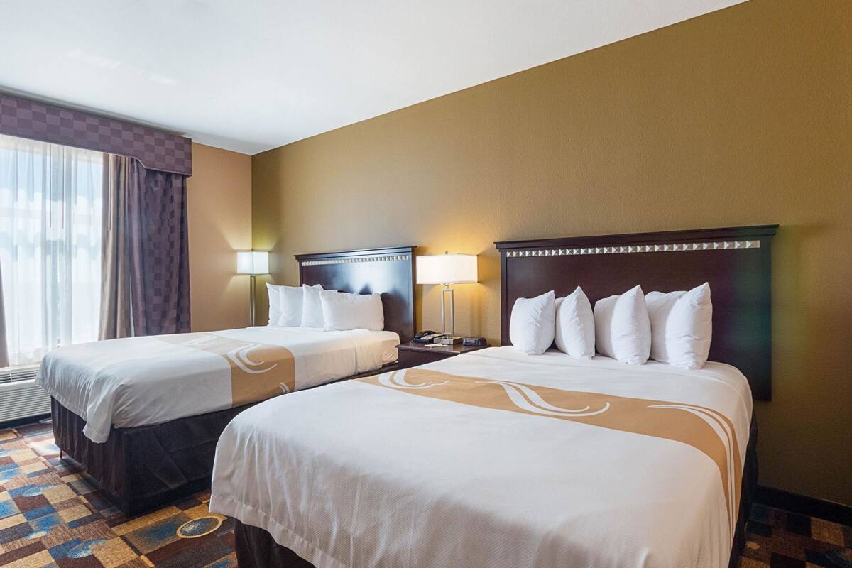 A hotel room with two beds, a nightstand, a lamp, and a window with curtains.