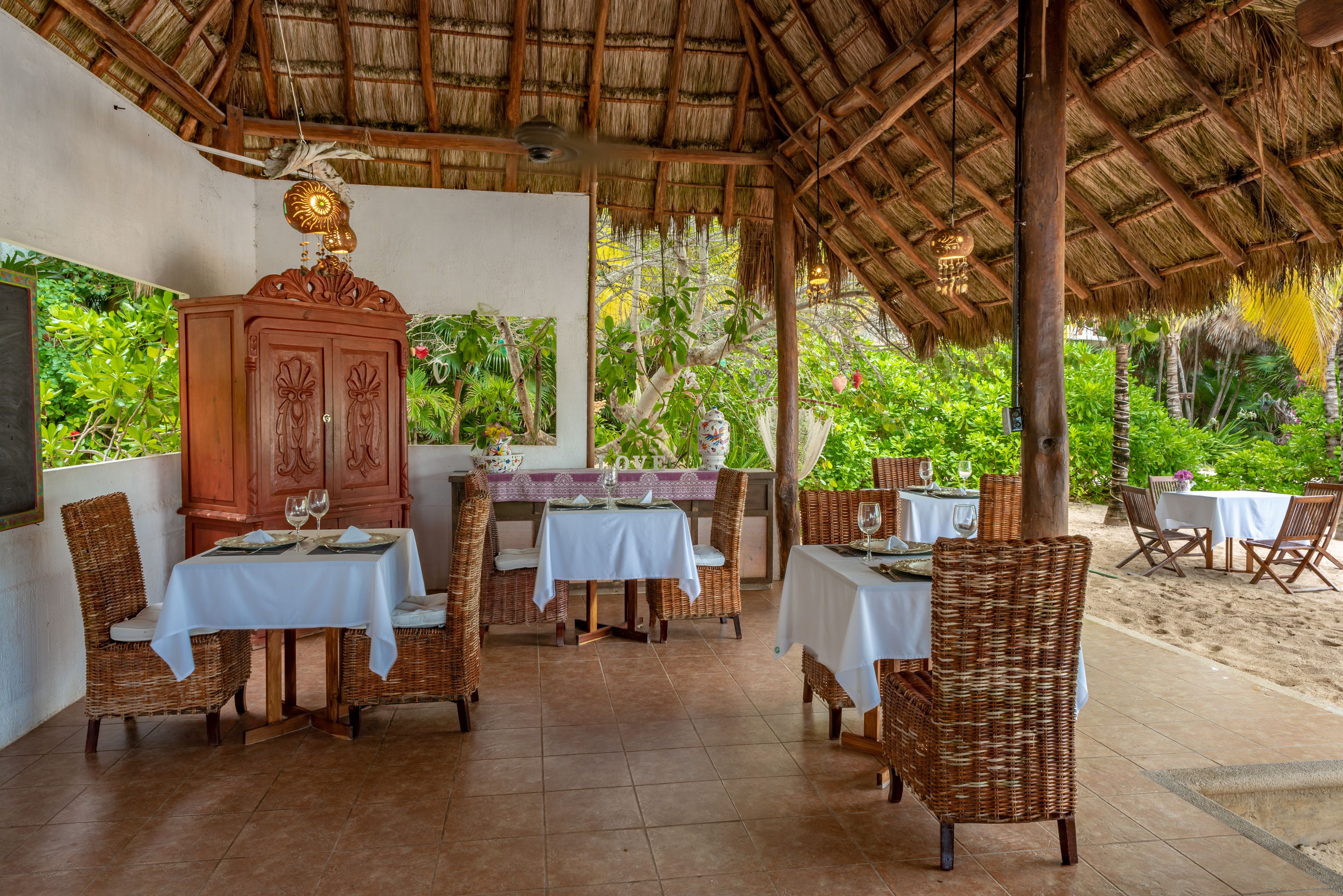 Un área de comedor al aire libre con sillas y mesas de mimbre, techo de paja y abundante vegetación de fondo.