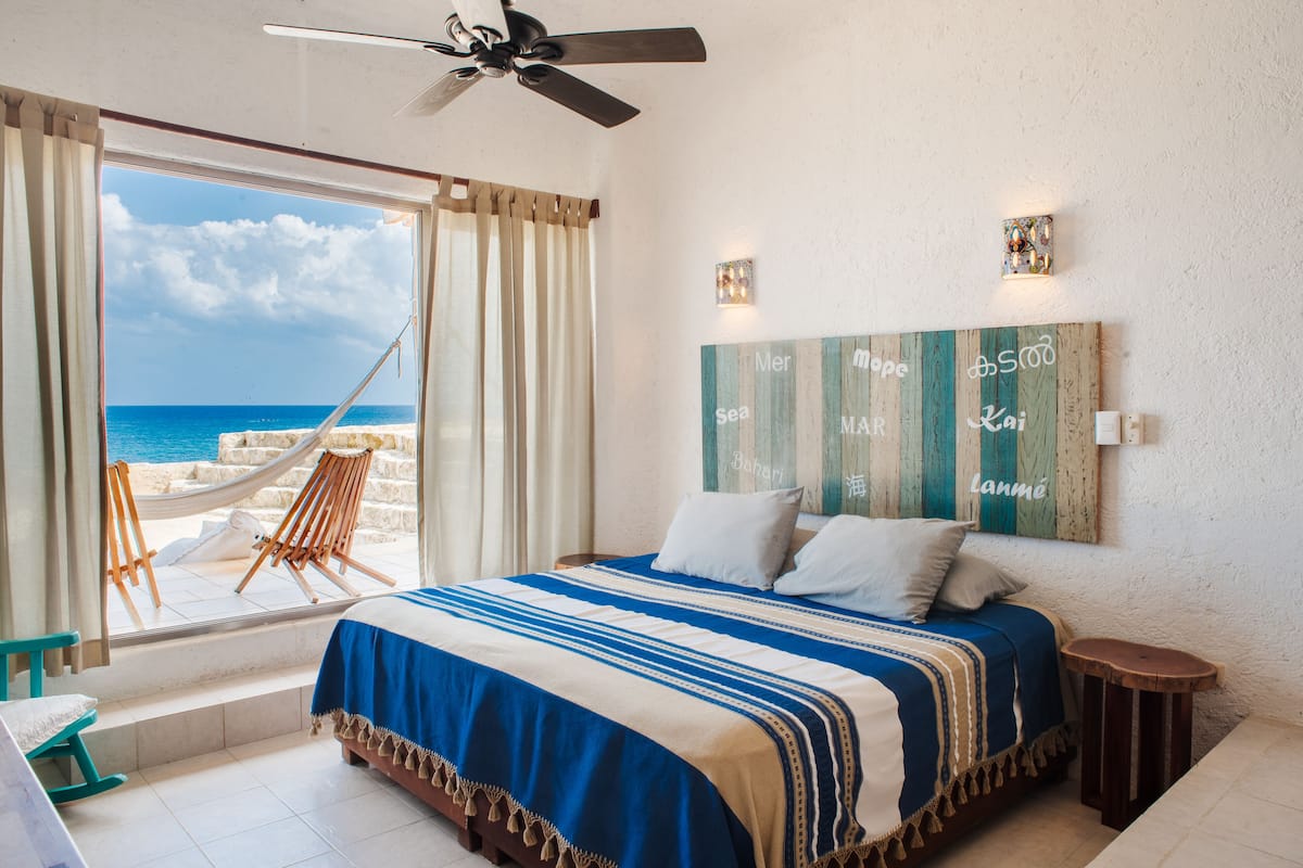 A bedroom with a large window, a bed with a striped bedspread, a hammock outside, and a wall with decorative text.