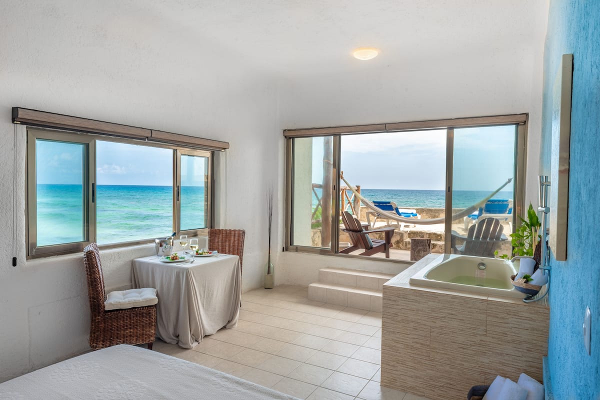 A room with a large window overlooking the sea, a bathtub, and a table with a chair.