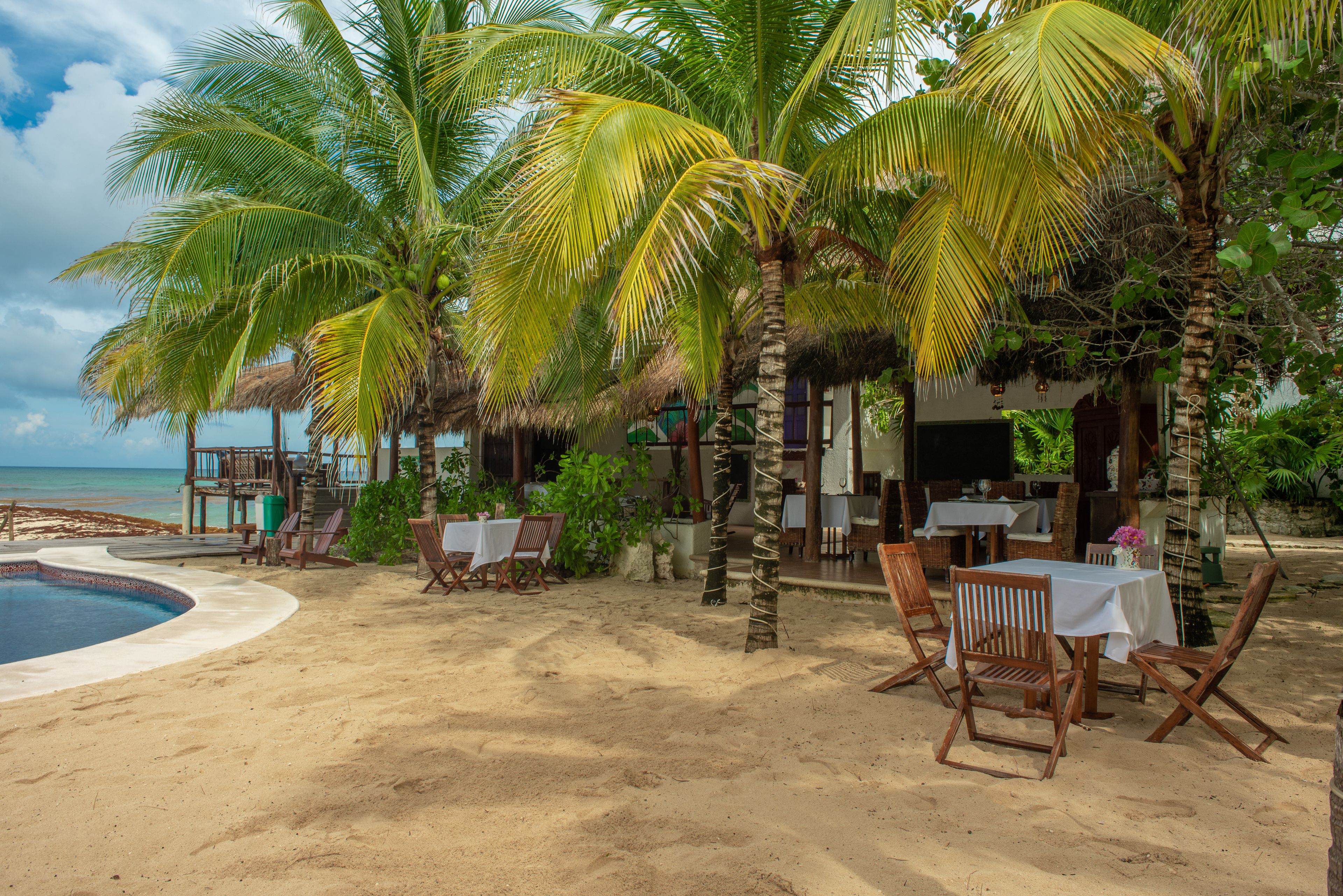 A beachside resort with a pool, wooden chairs, tables, and palm trees.