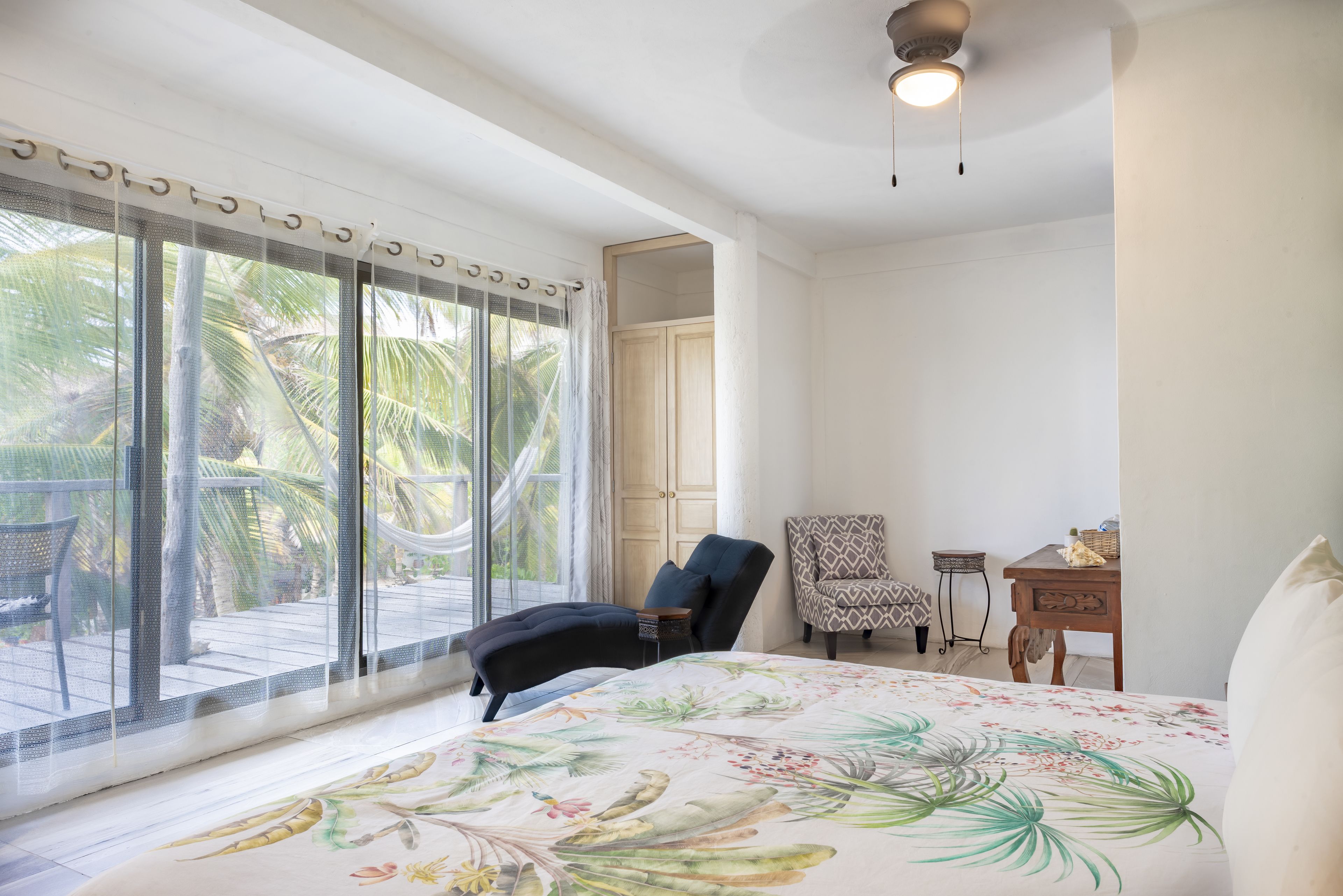 Un dormitorio con cama, una silla, una mesita y vistas a palmeras desde la ventana.